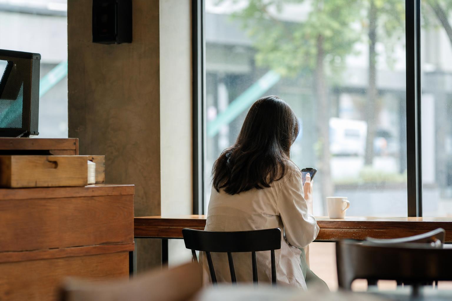 카페 창가에 앉아 스마트폰 화면을 들여다보는 사람의 뒷모습