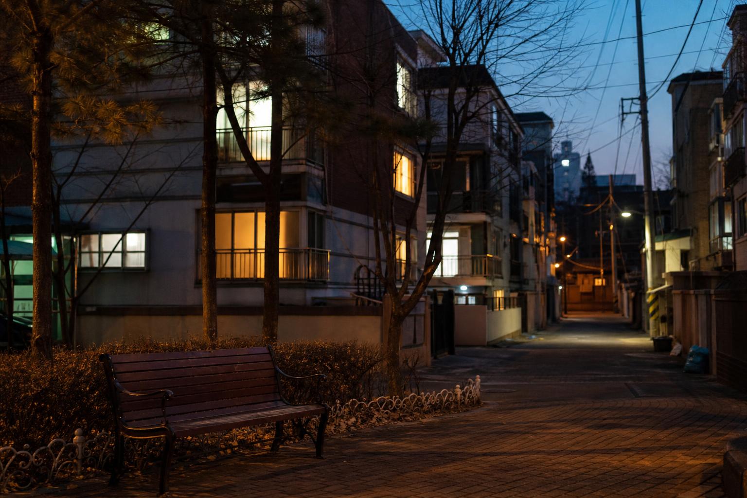 해질녘 조용한 한국 주택가 골목, 창문마다 따뜻한 불빛이 켜져 있고 빈 벤치가 앞에 놓여 있음