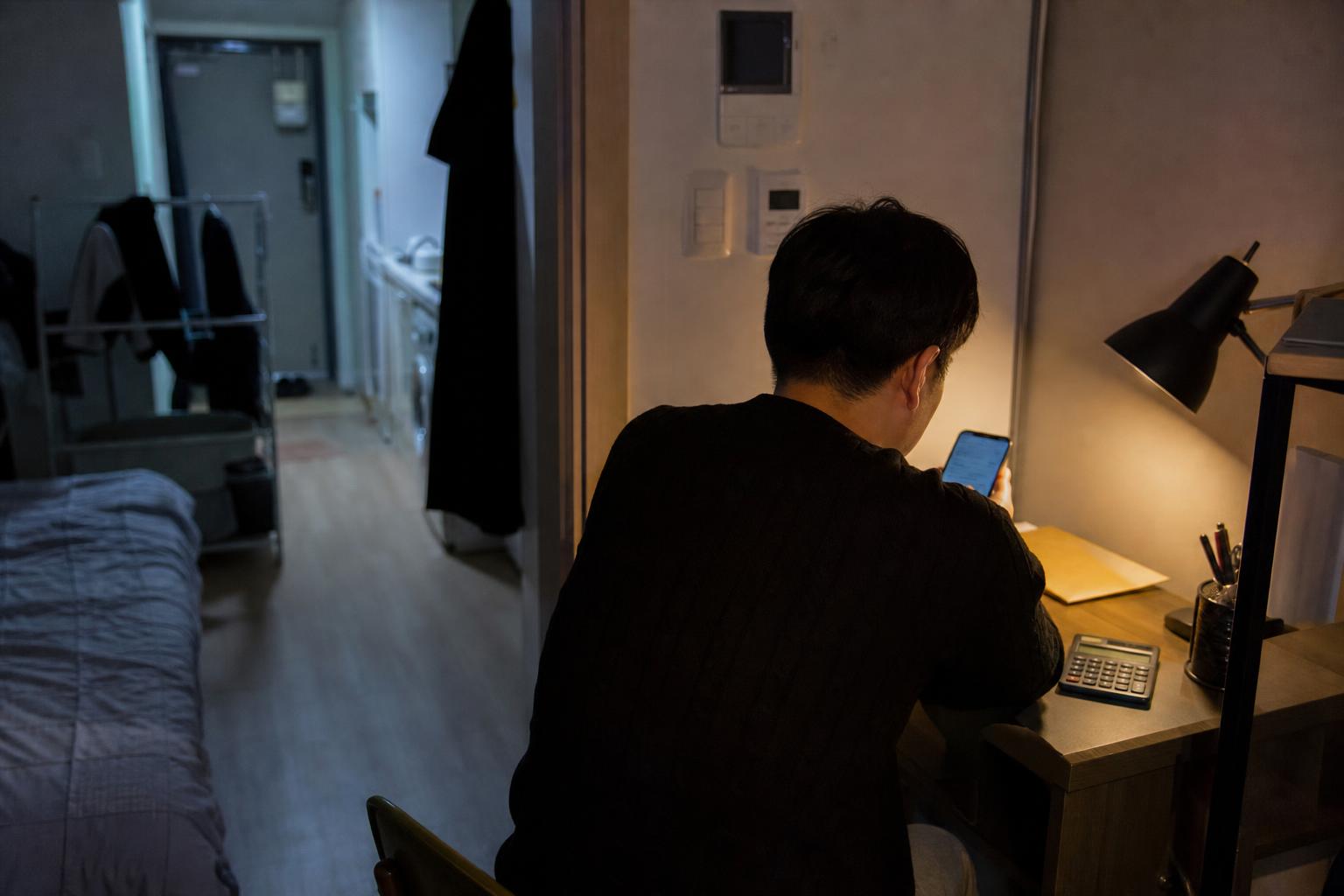한국의 원룸에서 책상에 앉아 휴대전화를 보는 사람의 뒷모습