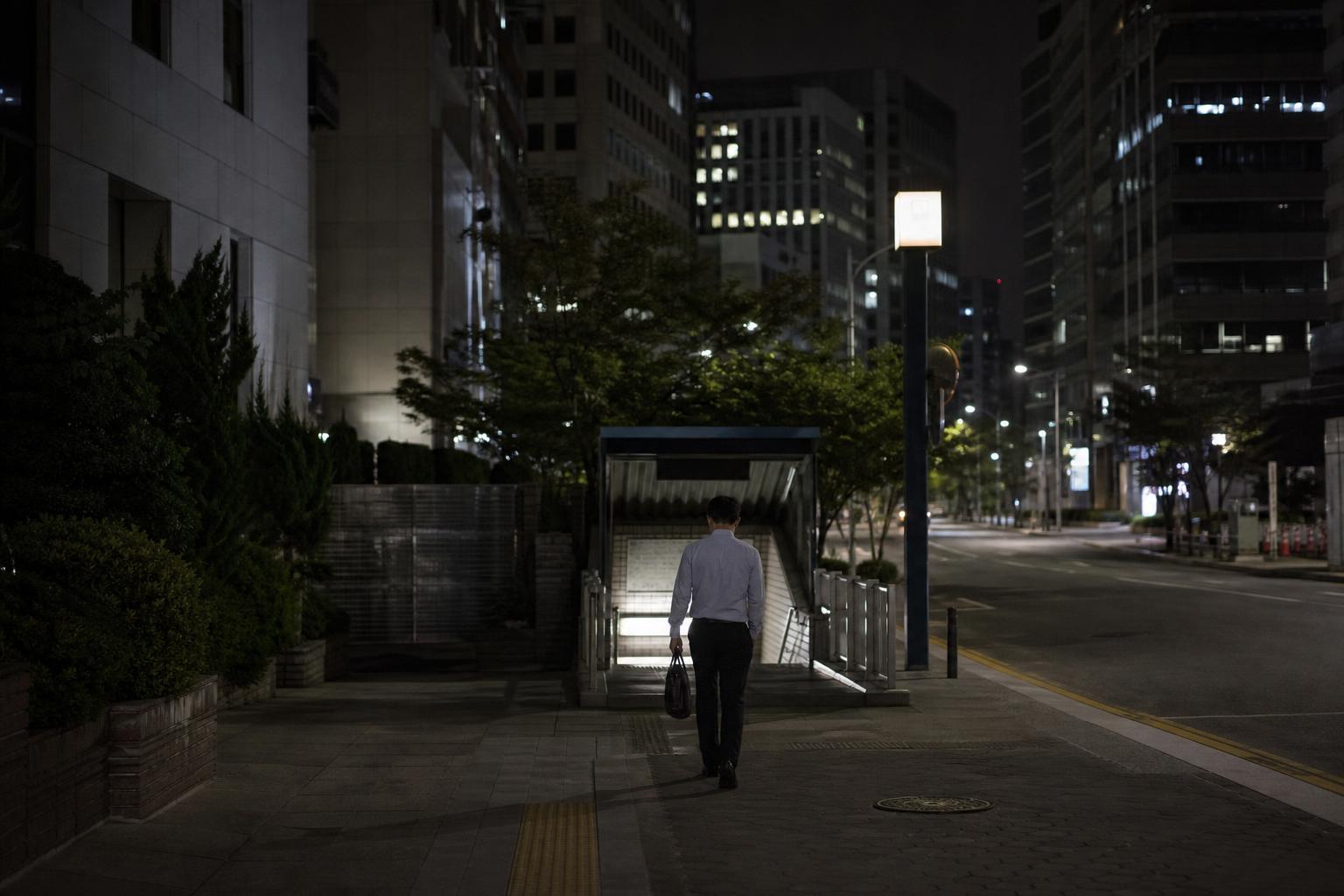 밤늦게 불이 켜진 한국의 사무실 건물 앞에서 지하철 입구로 걸어가는 직장인의 뒷모습